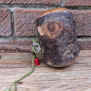 Könnte beinhalten: Ein handgefertigter Türstopper aus Holz mit natürlicher Holzmaserung. Der Türstopper hat eine zylindrische Form mit einem Knotendetail und einer kleinen roten Glocke, die mit einem grün-goldenen Seil befestigt ist. Der Hintergrund zeigt eine Backsteinmauer und eine Holzoberfläche.