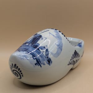 Könnte beinhalten: Weißer Keramik-Holzschuh mit blauen Windmühlen- und Blumenmustern. Der Schuh im holländischen Stil hat eine runde Spitze und eine flache Sohle. Die blauen Designs sind handbemalt.