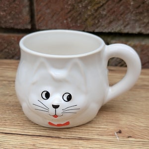 May include: White ceramic mug shaped like a cat's face with black eyes, whiskers, and a red bow tie.