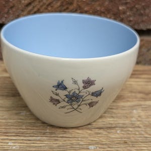 Op de afbeelding: Een witte keramische kom met een lichtblauwe binnenkant. De buitenkant heeft een bloemenpatroon in blauw, paars en bruin. De kom staat op een houten oppervlak.