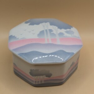 Könnte beinhalten: Achteckige Keramik-Schmuckschatulle mit einem pastellblauen, rosa und weißen Landschaftsdesign. Der Deckel zeigt eine stilisierte Darstellung von Bäumen und Wolken, während die Basis eine ähnliche Szene mit einer detaillierteren Ansicht der Bäume zeigt.