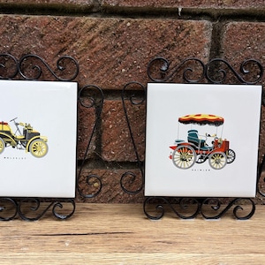 Könnte beinhalten: Zwei dekorative Kachelkunstwerke mit Illustrationen von Oldtimern. Jede weiße Kachel ist in einem schwarzen Schmiedeeisenrahmen eingefasst. Ein Auto ist gelb und schwarz, das andere rot und orange, beide mit gelben Rädern.