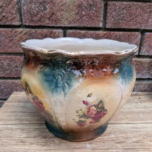 Puede incluir: Una maceta de cerámica con borde festoneado y acabado de esmalte craquelado. El exterior presenta un degradado en tonos crema, marrón y azul verdoso, decorado con motivos florales de rosas rosas y amarillas y patrones verticales en relieve.