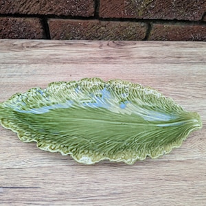 Puede incluir: Un plato de servir de cerámica verde con forma de hoja. El plato tiene una superficie texturizada y un borde ligeramente elevado.