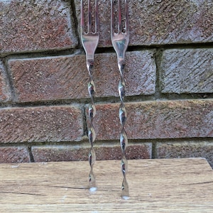 May include: Two silver-coloured metal cocktail forks featuring a three-tined head and a long, slender, spiraled twisted handle. The forks are constructed from a polished, reflective material and stand vertically, showcasing a uniform design.