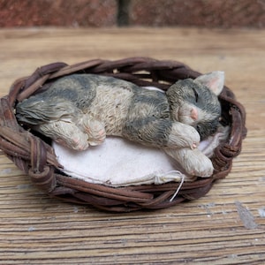 May include: A small figurine of a sleeping grey and white tabby cat curled up inside a miniature brown woven wicker basket. The cat rests on a small, off-white fabric cushion tucked into the base of the basket, with its eyes closed.