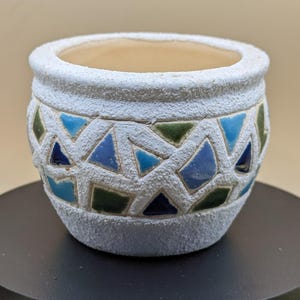 Op de afbeelding: Een kleine, ronde, witte keramische plantenbak met een getextureerd oppervlak. De plantenbak heeft een geometrisch ontwerp van blauwe en groene driehoeken. De binnenkant is lichtbeige. Ideaal voor kleine planten of vetplanten.