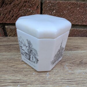 May include: A white ceramic trinket box with a hexagonal shape and a matching lid. The box features black and white illustrations of buildings. The box is sitting on a wooden surface.