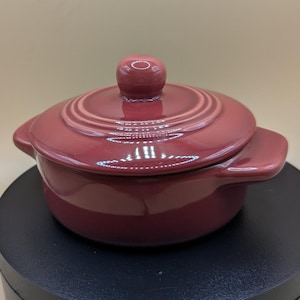 Op de afbeelding: Een bordeauxrode keramische ovenschotel met deksel. De schaal heeft een ronde body met twee zijhandvatten en een deksel met een ronde knop. De schaal staat op een zwart oppervlak.