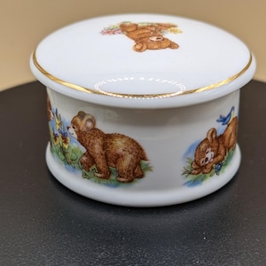 May include: A white ceramic trinket box with a lid, featuring illustrations of brown teddy bears with bluebirds and flowers. The box has a gold trim around the lid and base, and the bears are depicted in a playful, whimsical style.