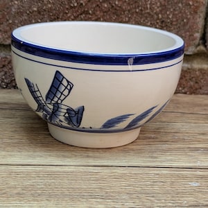May include: Cream ceramic bowl with a blue windmill design. The bowl has a blue rim and a footed base. The windmill design is repeated around the bowl. The bowl is on a wooden surface.