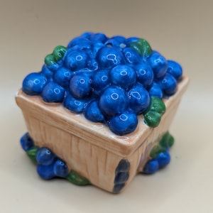 May include: A ceramic box shaped like a basket overflowing with glossy blue blueberries. The basket is tan with painted details, and green leaves and additional blueberries decorate the sides. A charming decorative piece.