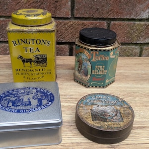 May include: A collection of four vintage metal tins including a yellow rectangular Ringtons Tea tin, a hexagonal Toffee tin with a black lid, a square silver Sarah Nelson's Grasmere Gingerbread tin, and a round Carr's Carpet Soap tin with a worn label.