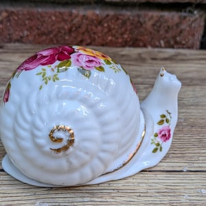 May include: A white ceramic snail figurine featuring a glossy finish and a spiraled shell decorated with vibrant red, pink, and yellow rose patterns. The snail has gold-painted accents on the shell center and edges, with small pink rose details on its neck.