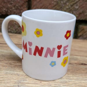 Puede incluir: Taza de cer&aacute;mica blanca con asa curva. La taza est&aacute; decorada con el nombre "MINNIE" en letras rosas y rojas, junto con dise&ntilde;os de flores y corazones coloridos. La taza est&aacute; dise&ntilde;ada para bebidas.