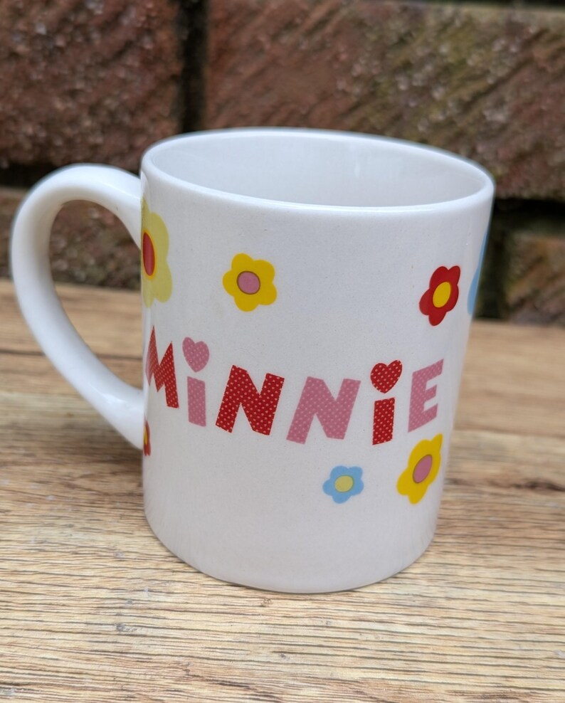 Puede incluir: Taza de cer&aacute;mica blanca con asa curva. La taza est&aacute; decorada con el nombre "MINNIE" en letras rosas y rojas, junto con dise&ntilde;os de flores y corazones coloridos. La taza est&aacute; dise&ntilde;ada para bebidas.