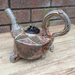 Könnte beinhalten: Eine handgefertigte Keramik-Teekanne mit einem einzigartigen Design. Die Teekanne hat eine braune und grüne Farbgebung mit einem dekorativen Griff und Ausguss. Der Deckel ist schwarz mit einem grünen Akzent. Die Teekanne ist etwa 18 cm lang und 15 cm hoch.