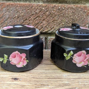 May include: Two black ceramic jars with lids. Each jar features a pink rose design with green leaves. The lids have a pink floral pattern and a gold rim. The jars are hexagonal in shape.