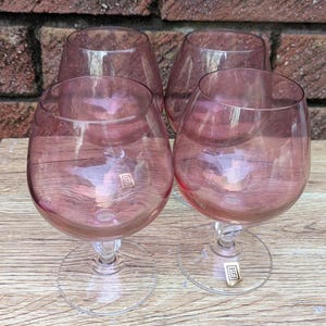 May include: Four stemmed brandy snifters with a pink hue. The glasses have a rounded bowl shape and sit on clear, detailed stems. The background features a brick wall and a wooden surface.