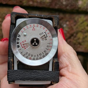 May include: Vintage light meter with a silver dial and black leather casing. The dial features ASA, DIN, and EV scales, along with aperture and shutter speed markings. A classic tool for photographers.