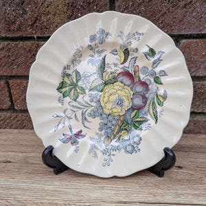 Puede incluir: Un plato de cerámica blanco con borde festoneado presenta un diseño floral colorido con frutas y hojas. El plato está sobre una superficie de madera.