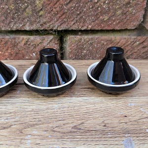 May include: Three black ceramic salt and pepper shakers with a white rim, arranged on a wooden surface. The shakers have a conical shape and a glossy finish, reflecting the light. The background features a brick wall.