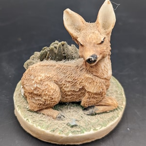 May include: A ceramic figurine of a fawn with a leafy base. The fawn is brown and white with a textured surface. The base is green and brown.