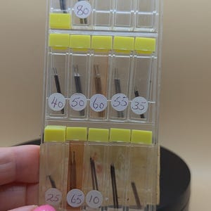 以下が含まれることがあります： 黄色い仕切りが付いた透明なプラスチック容器に、小さな濃い色の針が入っています。容器には80、40、50、60、55、35、25、65、10などの数字がラベル付けされています。容器は人が持っています。