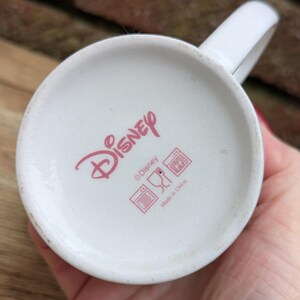 Puede incluir: Taza de cer&aacute;mica blanca con asa, con el logotipo de Disney en rosa en la base. La base de la taza tambi&eacute;n muestra s&iacute;mbolos de cuidado y el texto "Made in China".