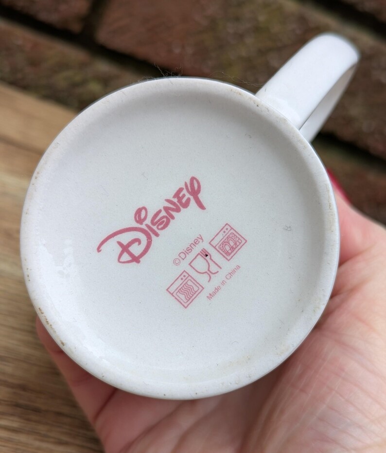 Puede incluir: Taza de cer&aacute;mica blanca con asa, con el logotipo de Disney en rosa en la base. La base de la taza tambi&eacute;n muestra s&iacute;mbolos de cuidado y el texto "Made in China".