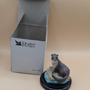 May include: A small, brown and white figurine of an otter sitting on a rock with blue water. The figurine is on a black base and comes in a Reader's Digest box. The box has the Reader's Digest logo and the number 021-510.