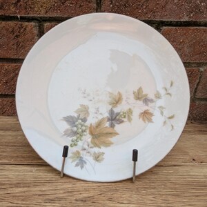 May include: A white ceramic plate with a floral design featuring green grapes and leaves in shades of green, brown, and gray. The plate is displayed on a metal stand against a brick background.