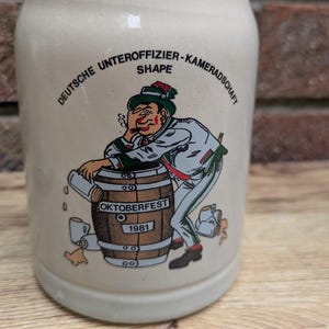 May include: A beige ceramic beer stein featuring a cartoon illustration of a man by a beer barrel. The barrel reads "Oktoberfest 1981". The stein has German text around the top.