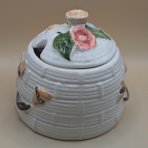 May include: White ceramic honey pot with a beehive design and a lid decorated with a pink flower and green leaves.