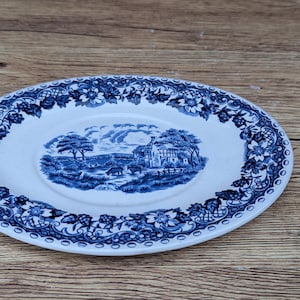 May include: A white ceramic plate featuring a blue transferware pattern. The center depicts a rural landscape with trees, a building, and grazing animals. The wide rim is decorated with a detailed floral and lattice border in blue.
