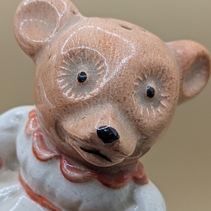 Könnte beinhalten: Vintage Keramik-Bärenfigur mit hellbraunem Kopf, schwarzen Augen und Nase. Der Bär trägt ein weißes Kleid mit rotem Kragen und Akzenten. Die Figur hat eine charmante, antike Ästhetik.