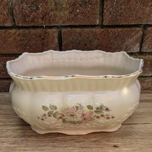 Puede incluir: Jardinera de cerámica color crema con borde festoneado y diseño floral. La jardinera presenta un ribete dorado y detalles en relieve. El patrón floral incluye flores rosas y amarillas con hojas verdes. Una pieza decorativa.