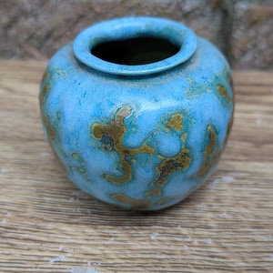 May include: A small, round ceramic vase with a wide opening and a thick, rolled rim. The exterior features a textured, mottled glaze in shades of bright turquoise blue, accented with irregular, organic patches of earthy brown and dark gold tones.