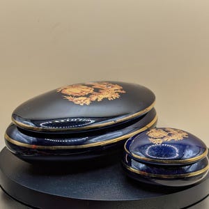 Puede incluir: Dos joyeros ovalados de cerámica azul marino con ribetes dorados y diseños florales. La caja más grande está apilada sobre la más pequeña. Las tapas presentan un diseño floral dorado.