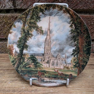 May include: A round decorative ceramic plate featuring a detailed landscape illustration of Salisbury Cathedral with a tall spire, surrounded by large trees, a grassy meadow, and several cows grazing near a small stream under a cloudy sky.