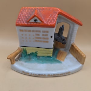 Könnte beinhalten: Eine weiße Keramikfigur eines Hauses mit rotem Dach und einem schwarzen Kaninchen, das aus dem Fenster schaut. Das Haus hat eine gelbe Tür und grünes Gras drumherum. Die Basis der Figur trägt die Aufschrift "The Lovers Barometer" und eine Liste von Sprüchen über die Liebe.