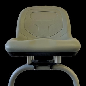 Puede incluir: Un asiento acolchado gris con una base negra y dos soportes plateados. El asiento tiene una superficie texturizada y un área rectangular elevada en la parte posterior.