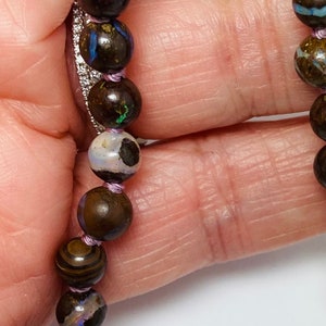 May include: A close-up of a necklace made of dark brown, black, and blue opal beads with a purple thread running through the beads.