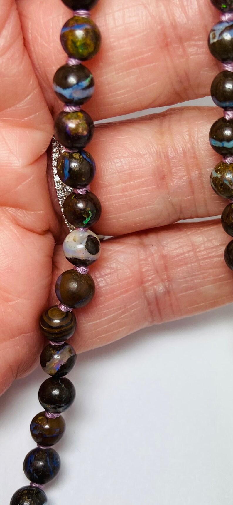 May include: A close-up of a necklace made of dark brown, black, and blue opal beads with a purple thread running through the beads.