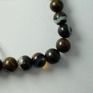 May include: A strand of dark brown opal beads with iridescent blue and white flashes. The beads are strung on a light purple cord.