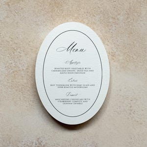 May include: An oval-shaped white menu card featuring black text. The menu lists an appetiser of roasted root vegetables, an entrée of beef tenderloin with demi-glaze and herb roasted mushrooms, and a dessert of mascarpone cheesecake with strawberry compote.