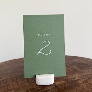 May include: A green table number card with the text "table no." and the number "2" in white. The card is held by a small, white rectangular stone. The card is on a dark wooden surface.