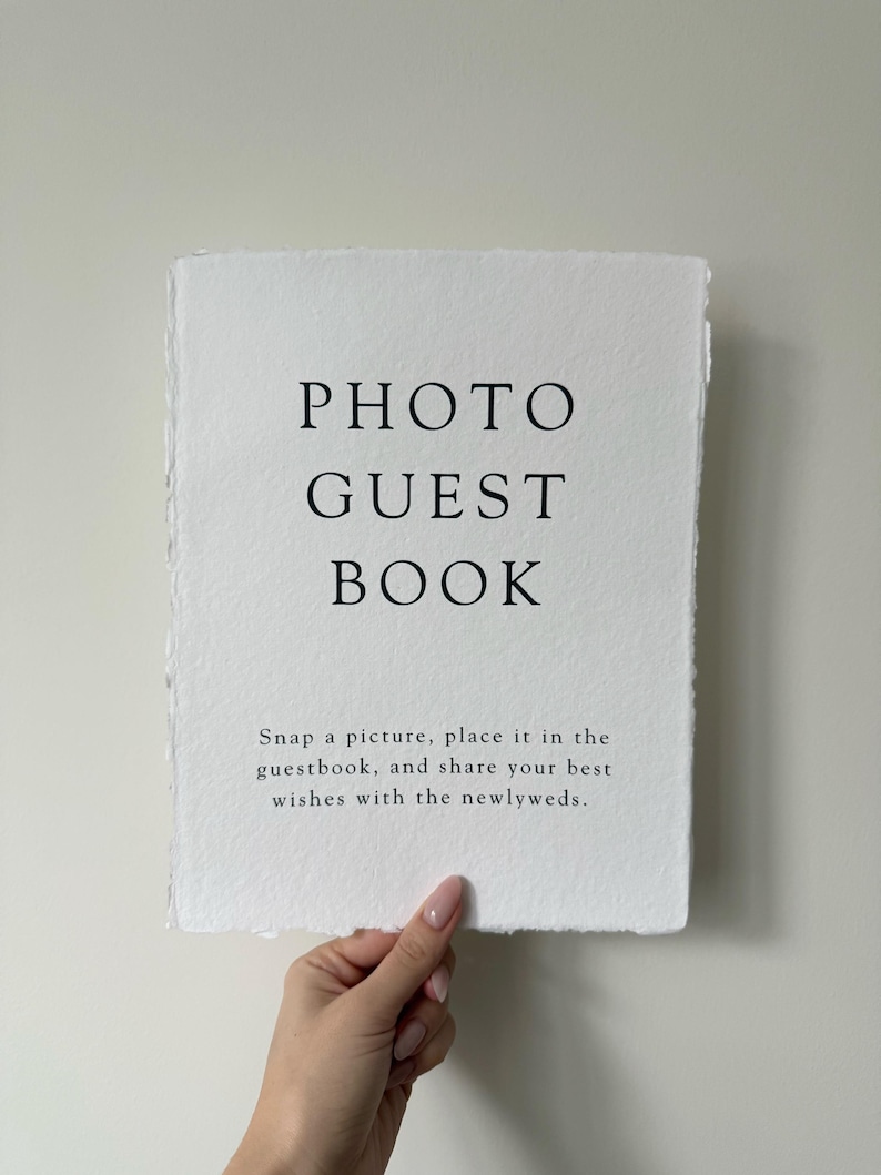 May include: White sign with black text that reads "PHOTO GUEST BOOK" and "Snap a picture, place it in the guestbook, and share your best wishes with the newlyweds."
