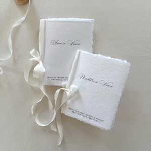 Può includere: Due libretti dei voti in carta fatta a mano bianca con bordi sfrangiati, legati con nastri di seta bianchi. Un libretto reca la scritta Sean's Vows e l'altro Maddie's Vows, entrambi con la data Friday the Twenty-Eighth of August Two Thousand Twenty-Six.