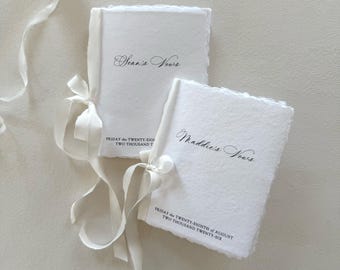 May include: Two white handmade paper vow books with deckled edges, tied with white silk ribbons. One book is inscribed with Sean's Vows and the other with Maddie's Vows, both featuring the date Friday the Twenty-Eighth of August Two Thousand Twenty-Six.